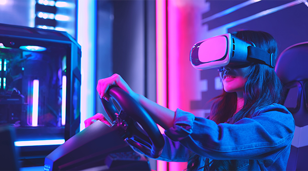 ALT Text: A woman with using a car simulator with a Virtual Reality headset on with a neon lights in the background