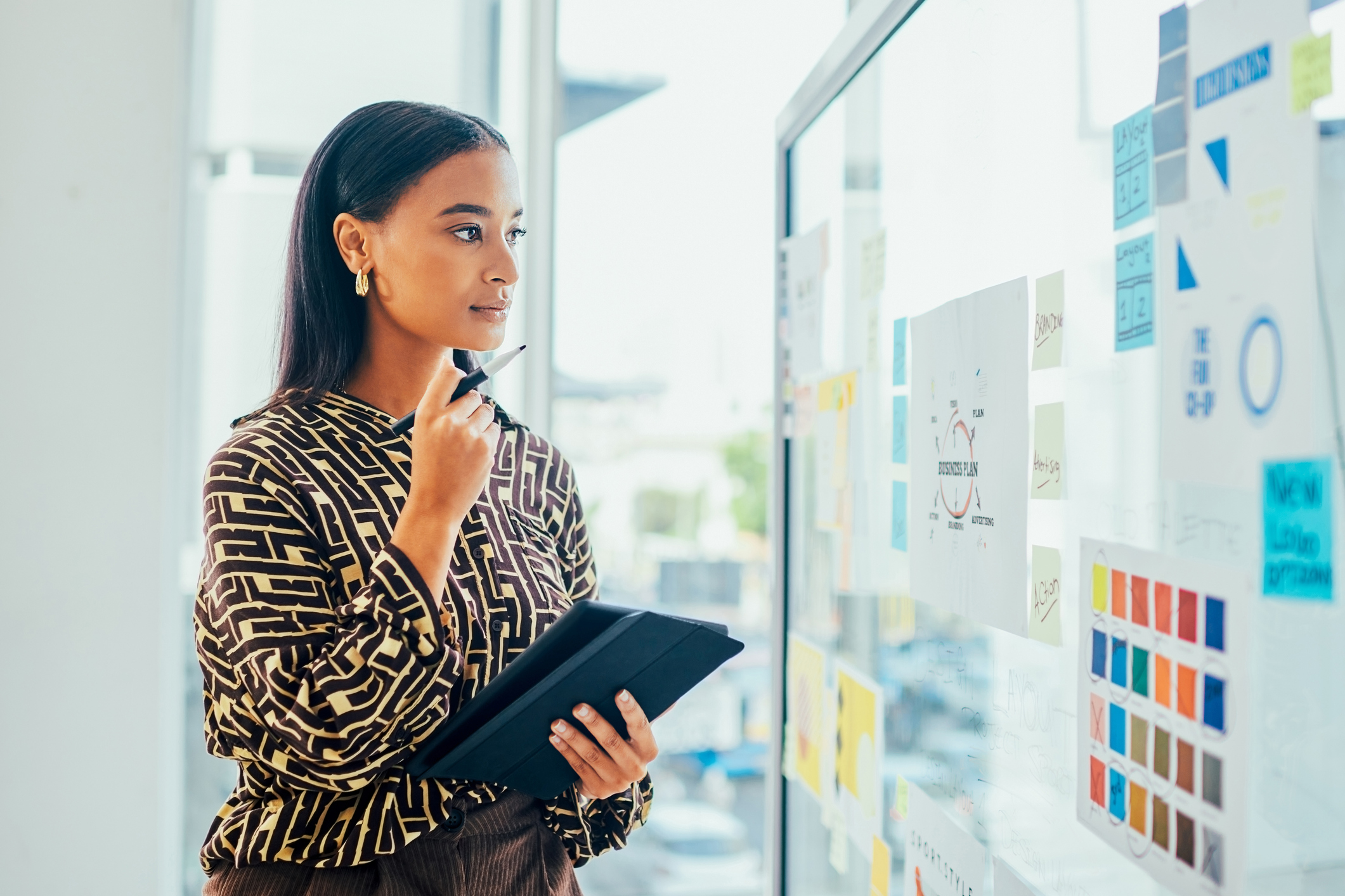 ALT Text: a woman dressed in cooperate attire looking at a whiteboard full of notes for a project and she is holding a tablet thinking